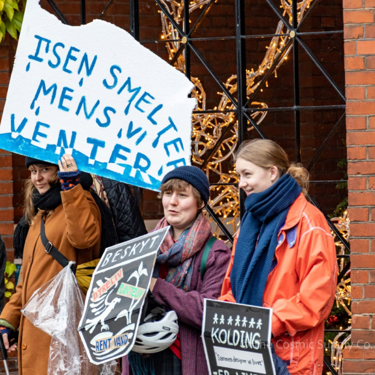 Three young woman wearing winter clothes and holding cardboard signs. The sign of the woman on the far left reads in Danish: the ice is melting while we are waiting
